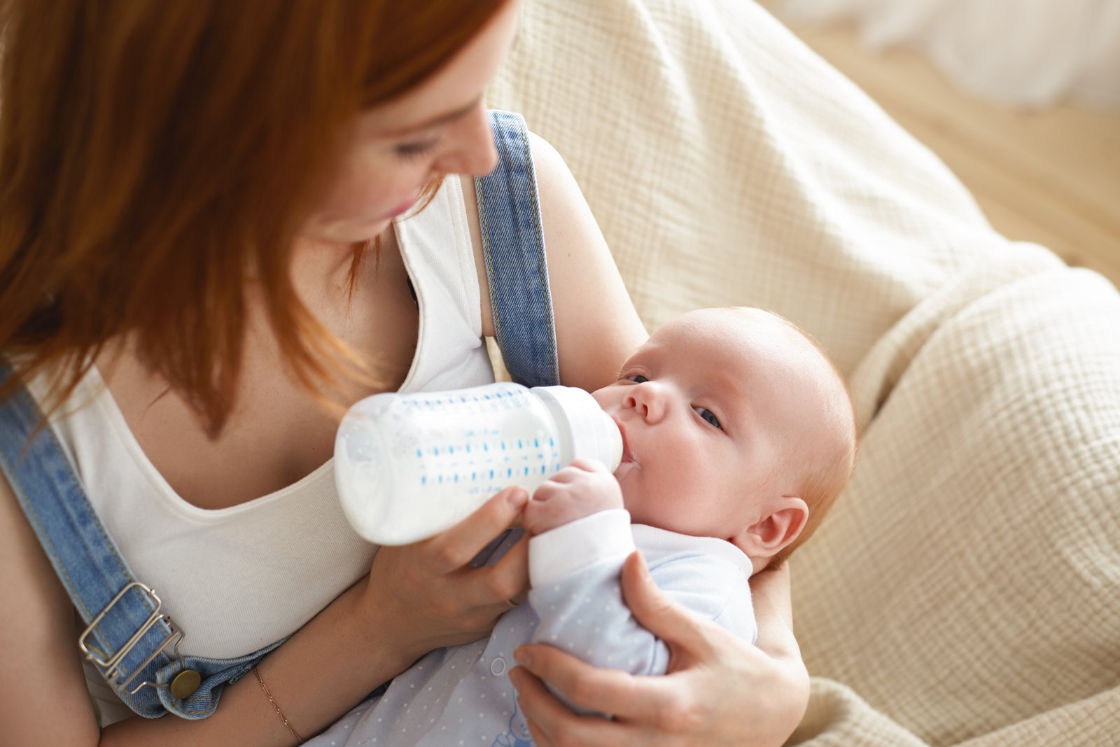 Portrait of cute infant resting in mother's arms, sucking on formula or expressed breast milk from glass nursing bottle. Tender young mom feeding her cute newborn child on couch in living room formula feed baby